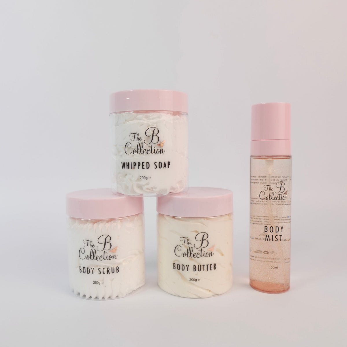 Four bath and body products from The B Collection arranged on a light gray background: two body scrubs in white jars with pink lids, one body butter jar in a white jar with a pink lid, and one body mist bottle in clear packaging with pink cap and pale pink liquid.