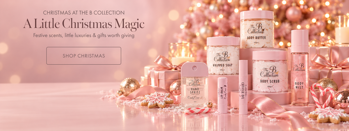 A festive collection of pink and gold holiday body care products from Collection, including body butter, body scrub, lip balm, and a soap bar, styled with wrapped gifts, candy canes, and warm bokeh lights in the background.